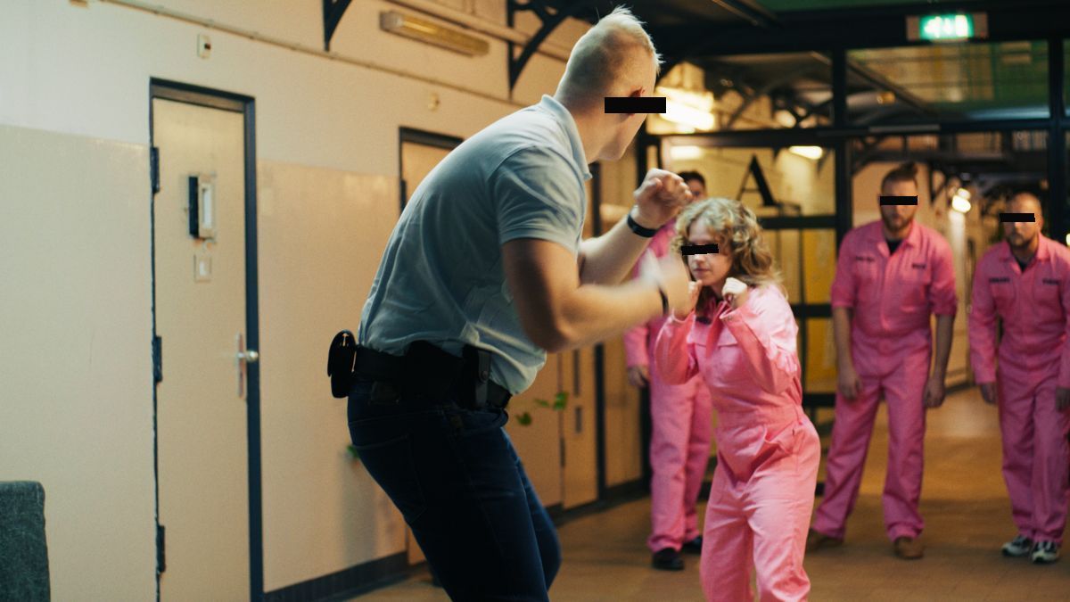 Mensen in roze overalls in een gevangenisgang, met zwarte balkjes over de ogen. Still uit een MediaTuig-commercial, 2024.