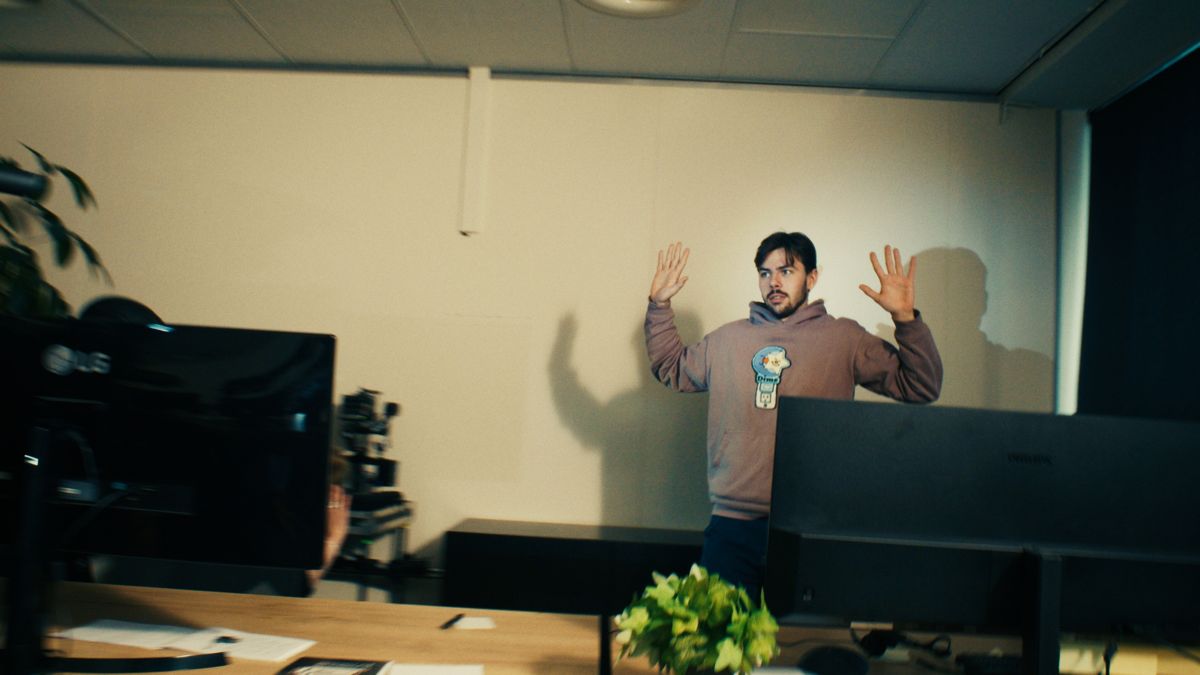 Een man in een hoodie staat met zijn handen omhoog tegen de muur van een kantoor. Still uit een MediaTuig-commercial, 2024.